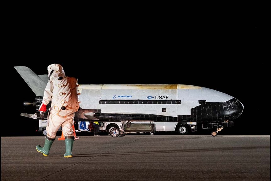 A man in protective gear walks toward Space Force's X-37B Space Plane sitting on a runway.