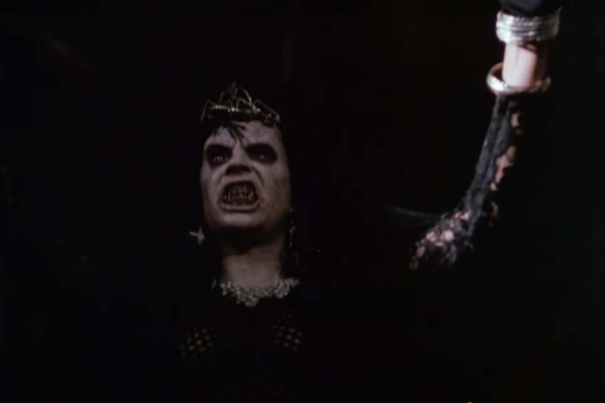 A demon dressed in all black and wearing a crown bares its monster teeth in Night of the Demons (1988).