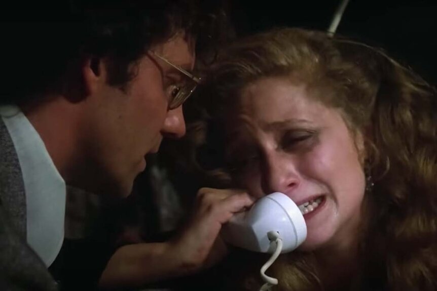 Jill Johnson (Carol Kane) (R) cries while speaking on a corded phone as a man watches in When A Stranger Calls (1979).