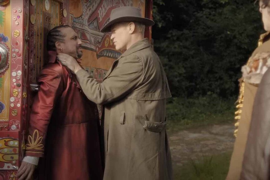 Wyatt Cain (Neal McDonough) wears a hat and trench while choking a man in a red coat while someone watches in Tin Man.