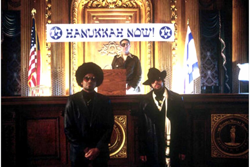 JJL Chief Bloomenbergensteinenthal (Peter Coyote) stands under a "Hanukkah Now" sign on a judge's stand while Mohammed Ali Paula Abdul Rahim (Mario Van Peebles) and Mordechai Jefferson Carver (Adam Goldberg) stand in front in The Hebrew Hammer (2003).