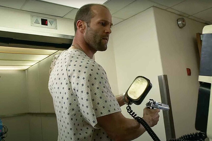 Chev Chelios (Jason Statham) wears a hospital gown and holds a defibrillator and gun in Crank (2006).
