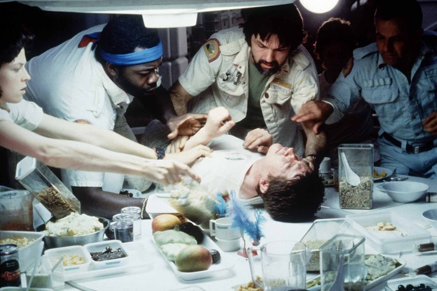 The cast of Alien (1979) tends to a crewmember on a dinner table.