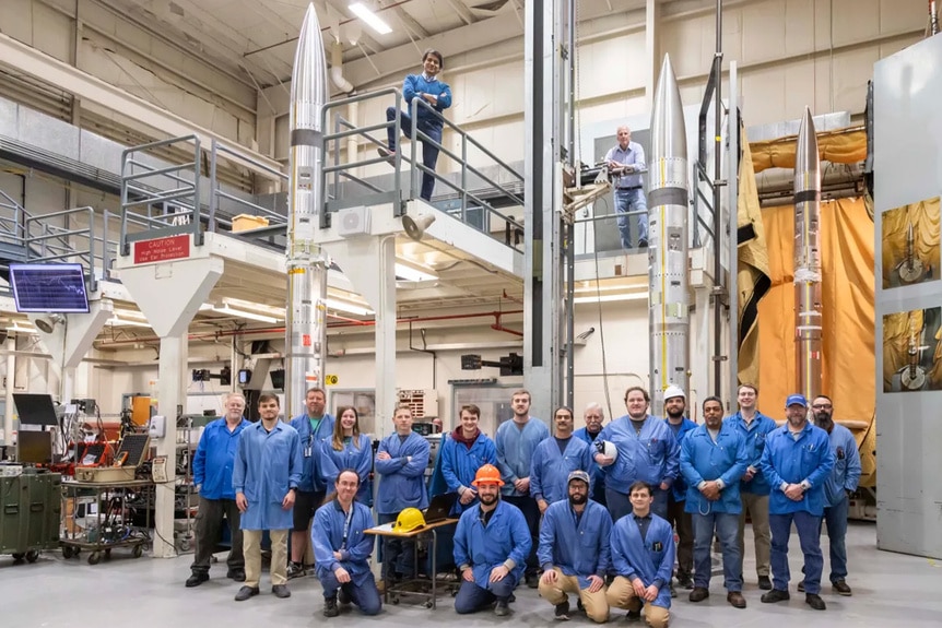 The NASA support team poses in blue shirts next to three APEP sounding rockets.