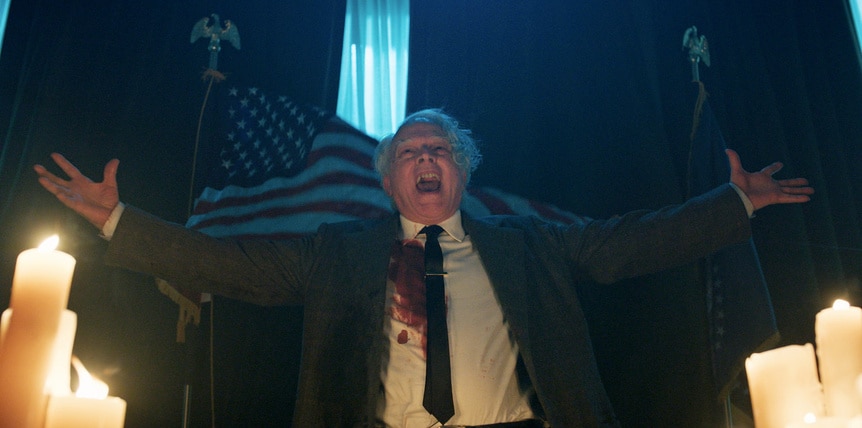 Charles Lee Ray cackles near an American flag and candles in Chucky Episode 307.