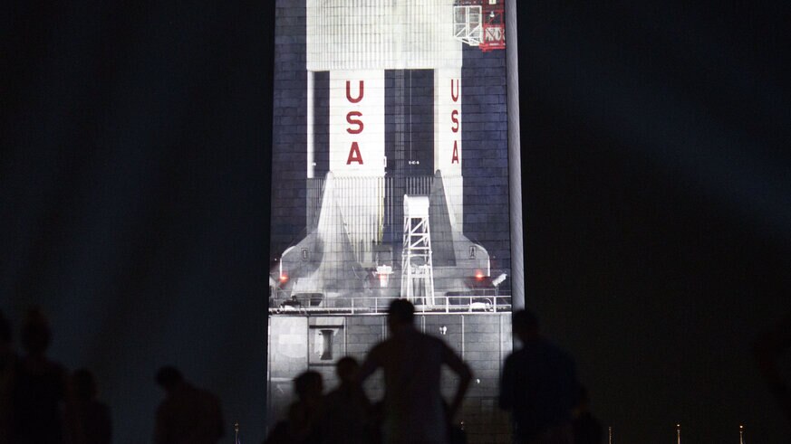 The Saturn V rocket projected at night against the Washington Monument
