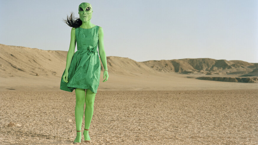 Extraterrestrial wearing green dress standing in desert.